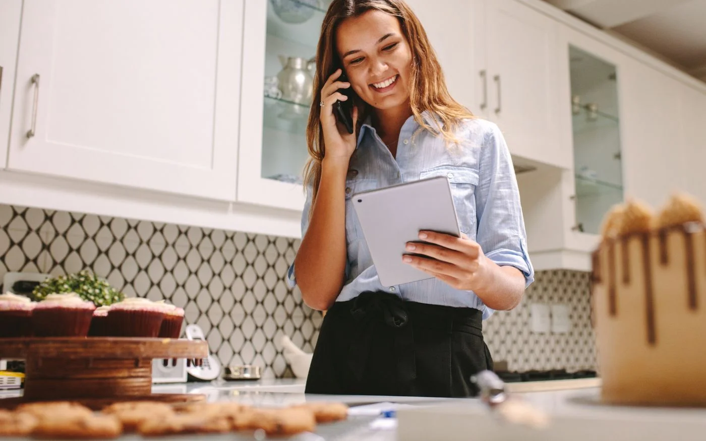 A woman standing in front of a kitchen full of cupcakes, engaged in a phone conversation.